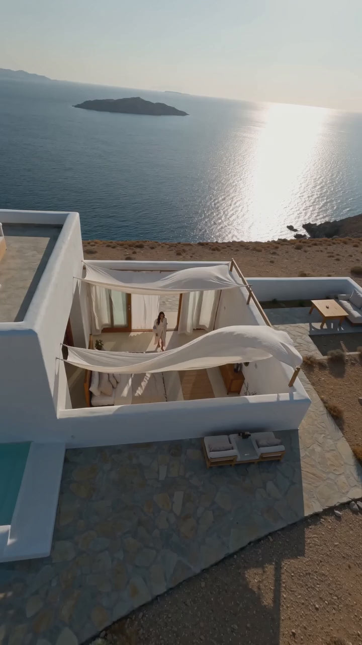 Woman wearing a long cream textured cotton kimono robe with blue evil eye motifs, wide sleeves, fringe trim and a matching waist belt while walking through a sunlit Mediterranean-style seaside home. The video shows close-up details of the blue eye design and natural woven fabric, followed by the full robe silhouette and relaxed movement against an ocean view.