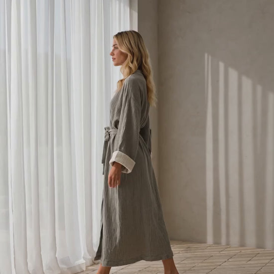 Woman wearing the soft gray Zen Turkish cotton muslin kimono robe by a sunlit window, highlighting its textured gauze fabric, ivory cuffs and relaxed silhouette.
