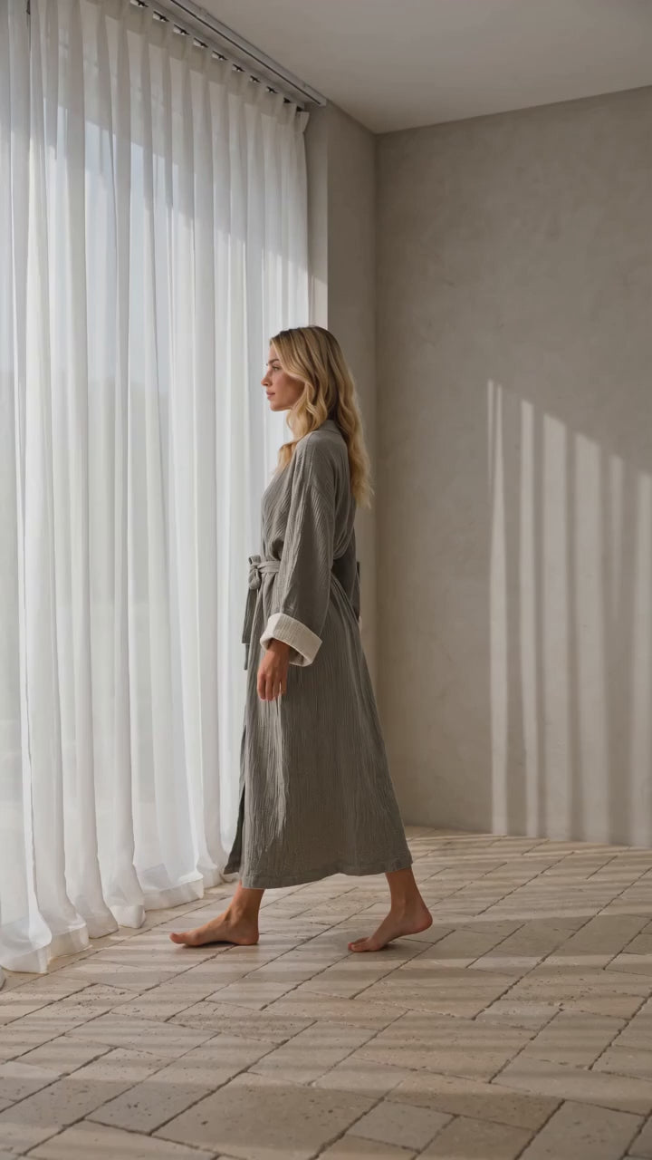 Woman wearing the soft gray Zen Turkish cotton muslin kimono robe by a sunlit window, highlighting its textured gauze fabric, ivory cuffs and relaxed silhouette.