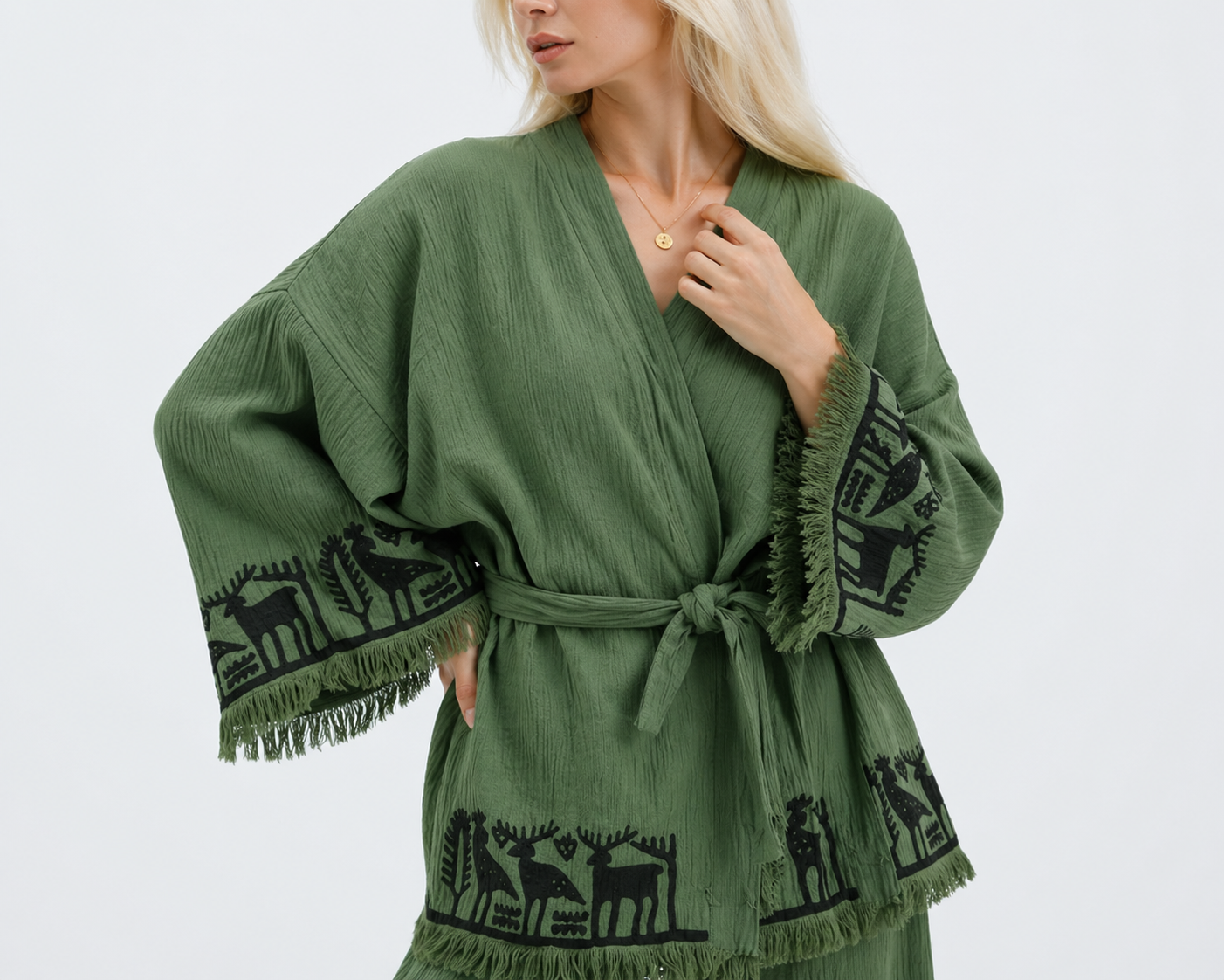 Close-up of a sage green textured cotton kimono jacket showing black Anatolian-style deer, bird, and tree motifs, a matching waist tie, wide sleeves, and hand-finished fringe trim.