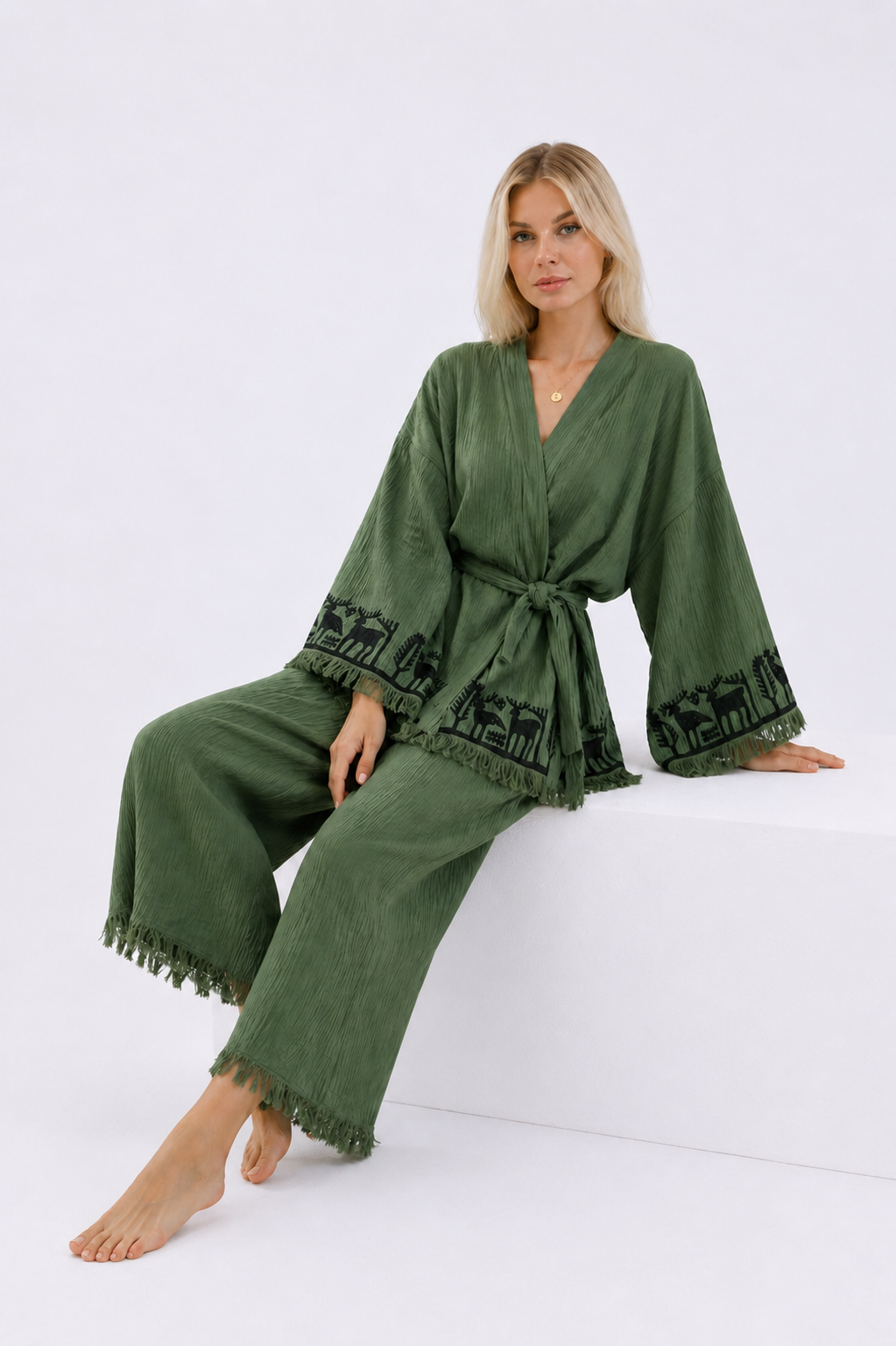 Seated model wearing a sage green textured cotton kimono and wide-leg pants set with black Anatolian animal and tree motifs, a belted waist, wide fringed sleeves, and relaxed cropped pants.