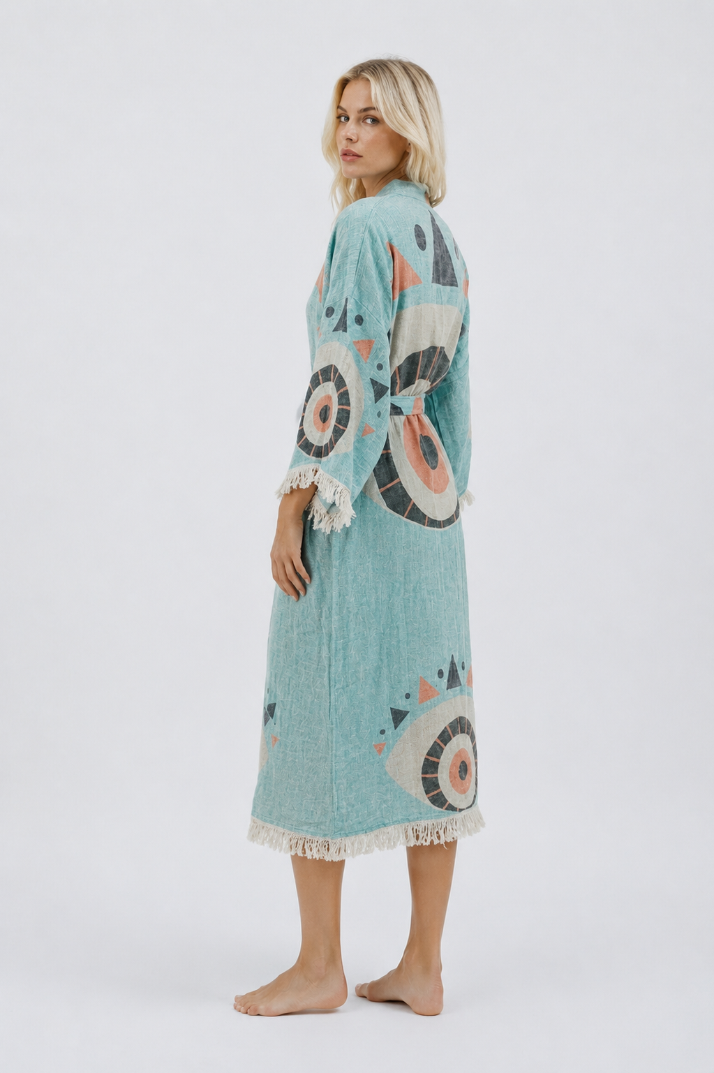Side-back view of a turquoise jacquard cotton kimono robe showing the relaxed long silhouette, oversized evil eye motifs, matching waist belt, textured fabric, and cream fringe edging.