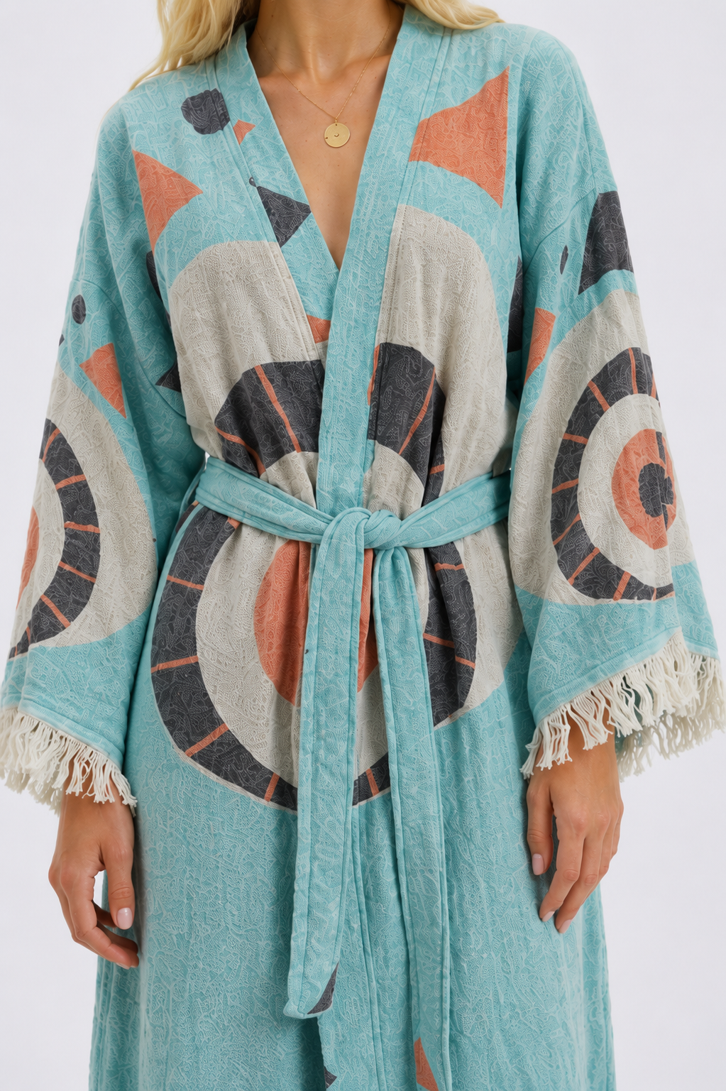Close-up of a turquoise jacquard cotton kimono robe showing the woven fabric texture, coral and charcoal evil eye pattern, matching tie belt, wide sleeves, and hand-finished cream fringe.