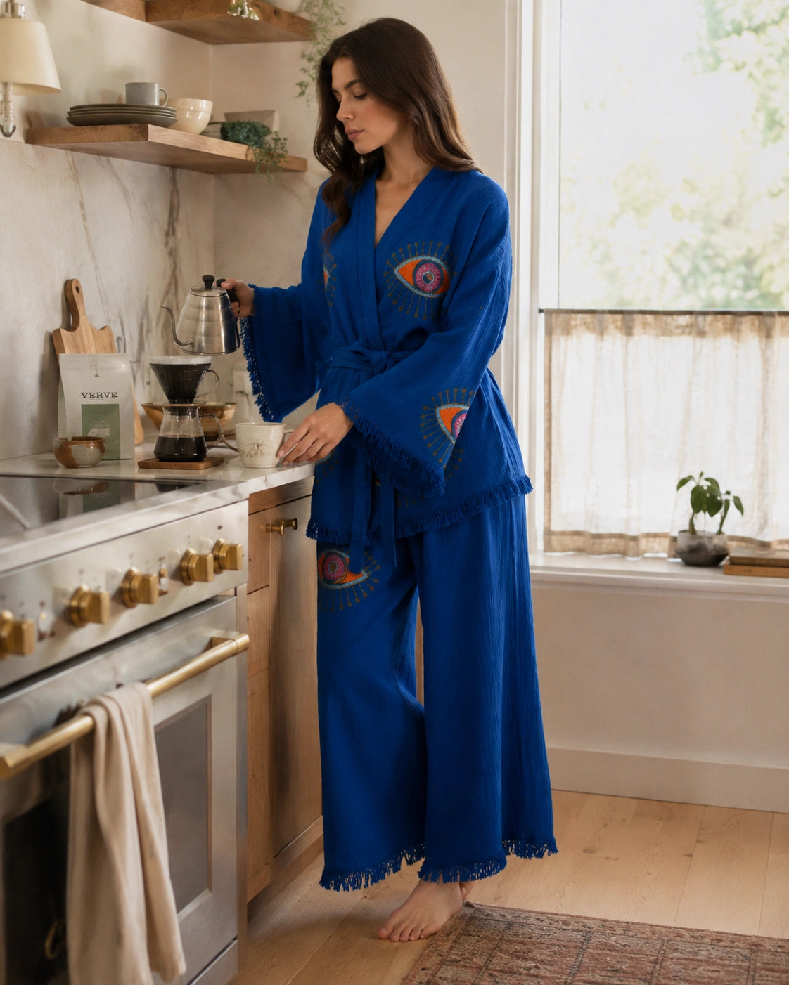 Woman wearing the Zaria Blue Protection Kimono Robe Set while making coffee at home, showing the cobalt blue textured fabric, wide-leg pants, fringe trim and colorful protective eye motifs.